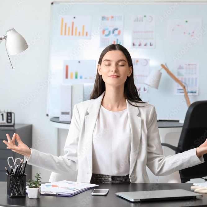 Eine Frau sitzt mit geschlossenen Augen und den Armen und Fingern im Meditationsgriff an einem Schreibtisch in einem Büro.