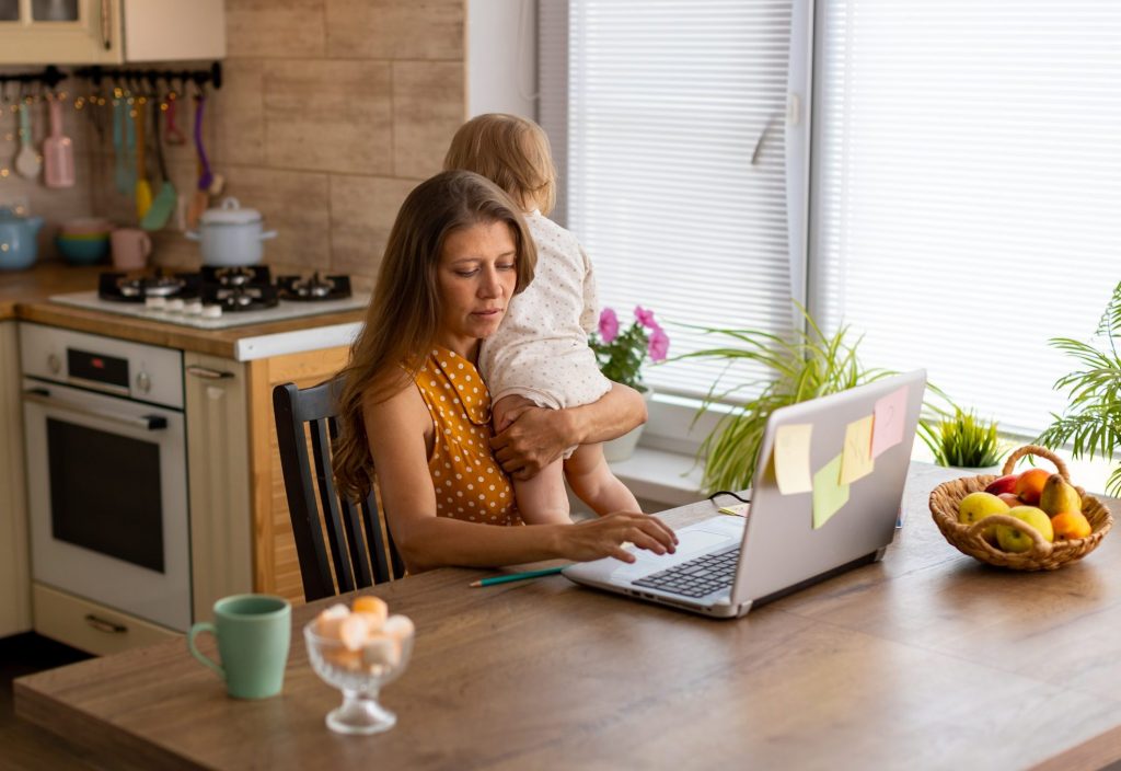 Eine Frau sitzt an einem Küchentisch und tippt mit der rechten Hand an einem Laptop, im linken Arm hält sie ein Baby.