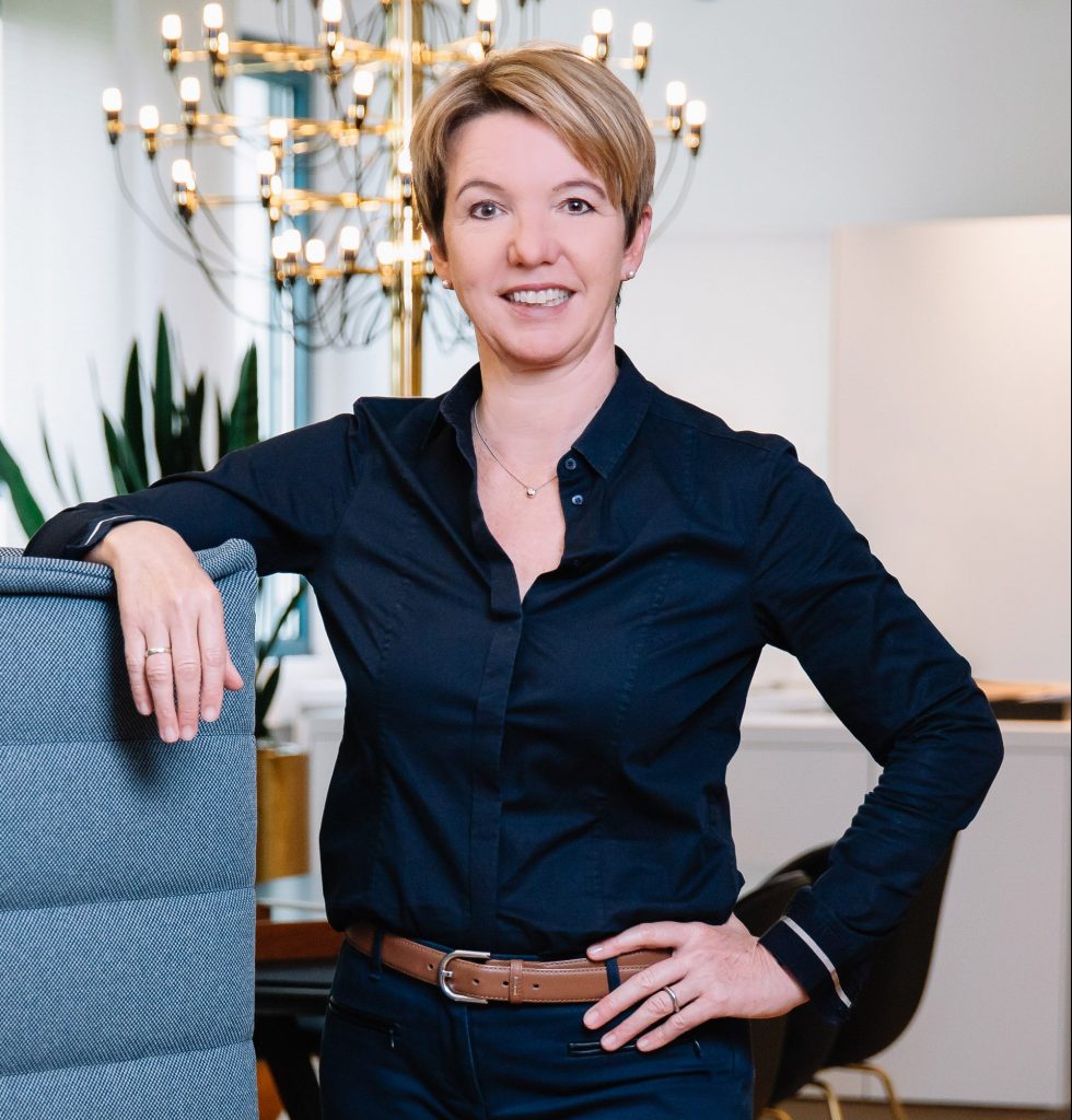 Ergonomieberaterin Susanne Weber steht dynamisch an einem Bürostuhl und guckt freundlich in die Kamera.