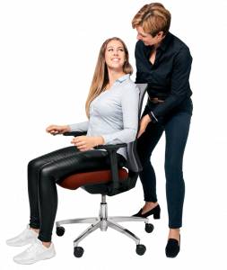 Eine junge Frau sitzt auf einem Bürostuhl, hinter ihr steht Ergonomie-Beraterin Susanne Weber und zeigt ihr die korrekte Sitzhaltung.
