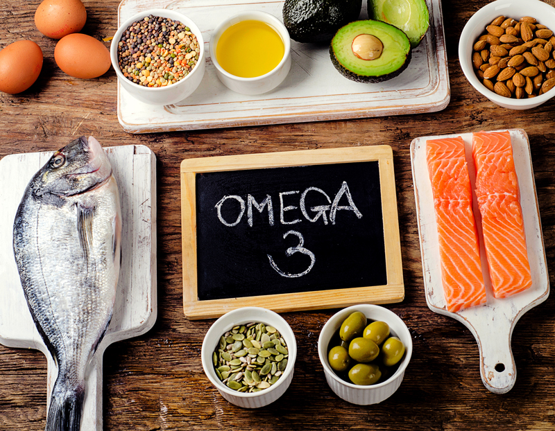 Ausschnitt von einem Tisch mit einer Tafel, auf der OMEGA 3 steht mit verschiedenen Lebensmitteln drumherum. Eier, Hülsenfrüchte, Öl, Avocados, Mandeln, Lachs, Oliven, Kerne, ganze Dorade.